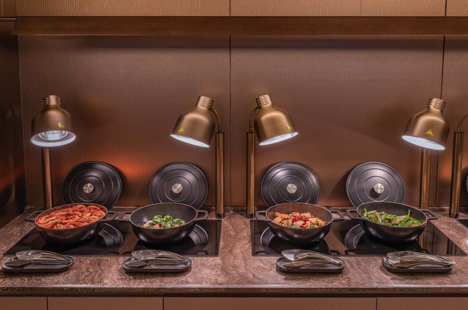 A kitchen with four induction cooktops, each with a pan of food, and overhead lighting.
