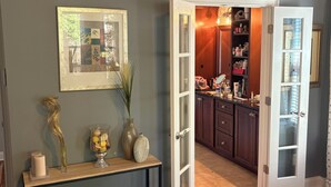 A bedroom with a wooden floor, a bedside table, a nightstand, a bed, a doorway leading to a bathroom, and a hallway. - Main level access = 2 BR, 2 private baths, and much more. Wheelchair accessible. (Braselton)
