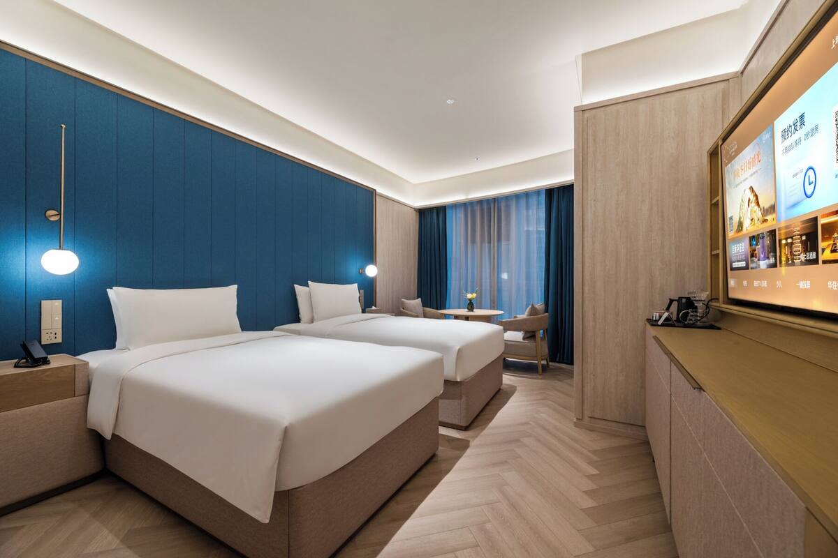 A hotel room with two beds, a desk, a chair, a TV, and a minibar.