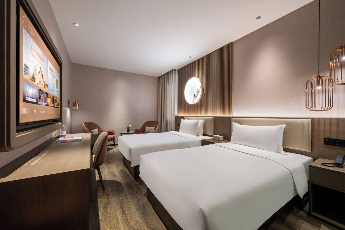 A hotel room with two beds, a desk, a chair, a TV, and a modern light fixture.