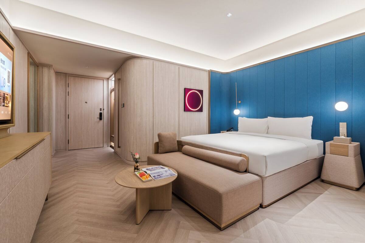 A modern hotel room with a large bed, a small seating area, a desk, and a wall-mounted TV.