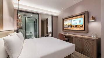 Ein Hotelzimmer mit Bett, Schreibtisch, Fernseher und einer Dusche mit Glastrennwand.