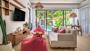 Una sala de estar moderna con un sofá, sillones tipo puff rojos, un televisor montado en la pared y un ventanal con vistas a un jardín y una piscina.