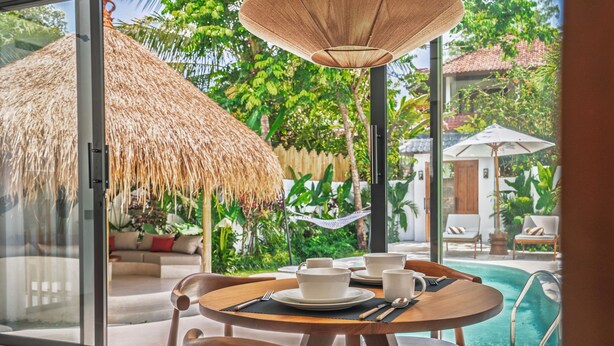 Un comedor con una mesa dispuesta para dos, una sombrilla de paja y vistas a una piscina y vegetación exuberante.