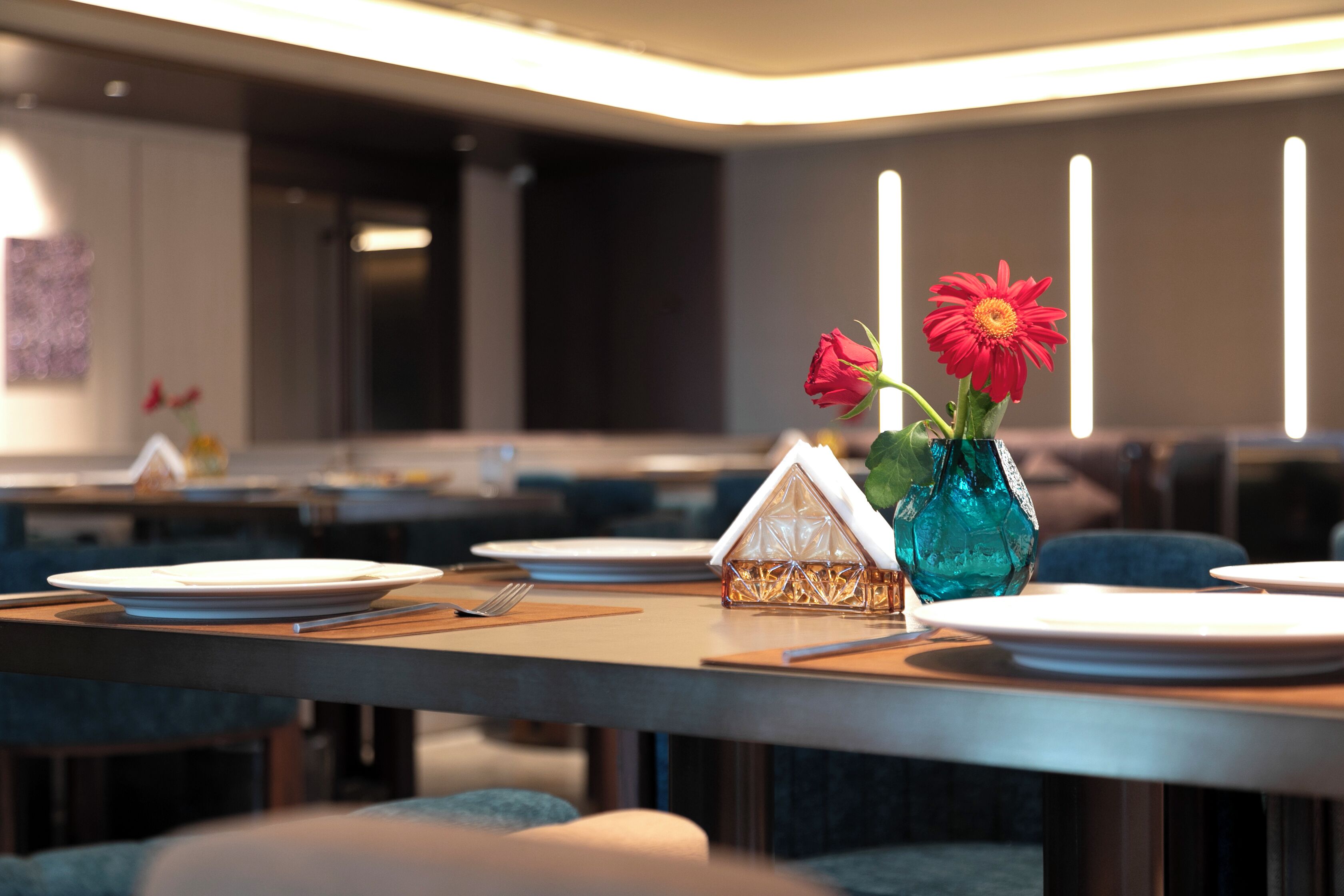 A dining area with tables set for two, a blue vase with red flowers, and a small house-shaped salt and pepper shaker.