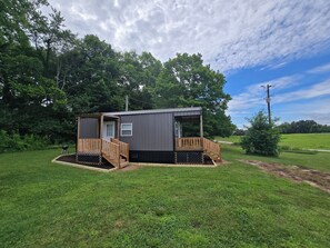 A small, single-story cabin with a wooden deck and a covered porch, surrounded by lush greenery. - Charming Tiny House in Country (Adairville)