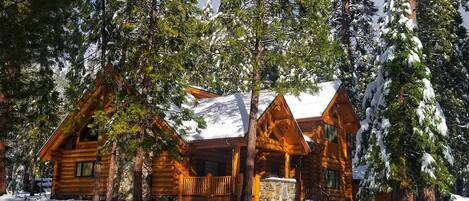 A log cabin with a stone foundation, surrounded by snow-covered trees.