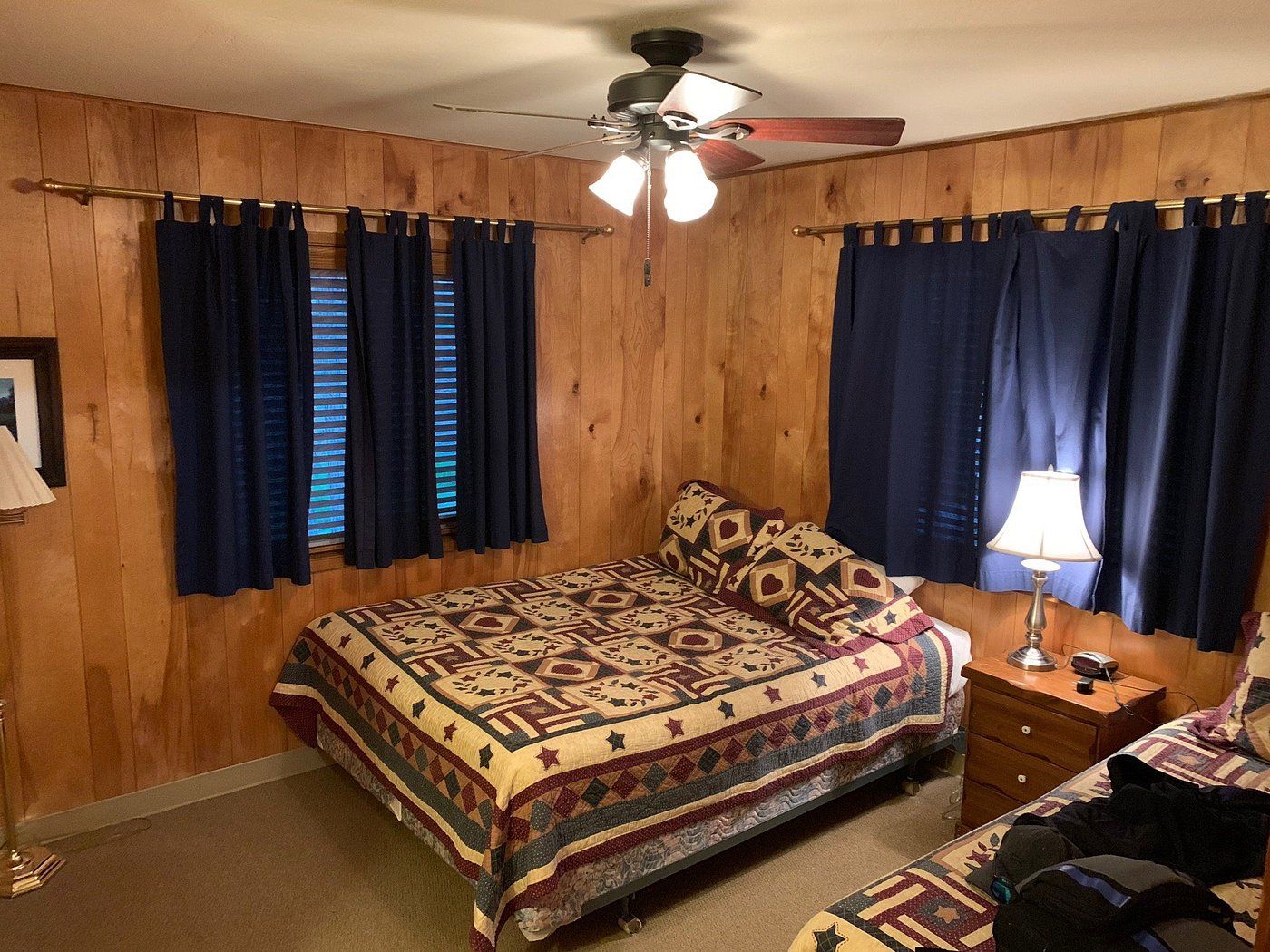 A bedroom with a wooden paneled wall, a bed with a patterned bedspread, a nightstand with a lamp, and a ceiling fan.