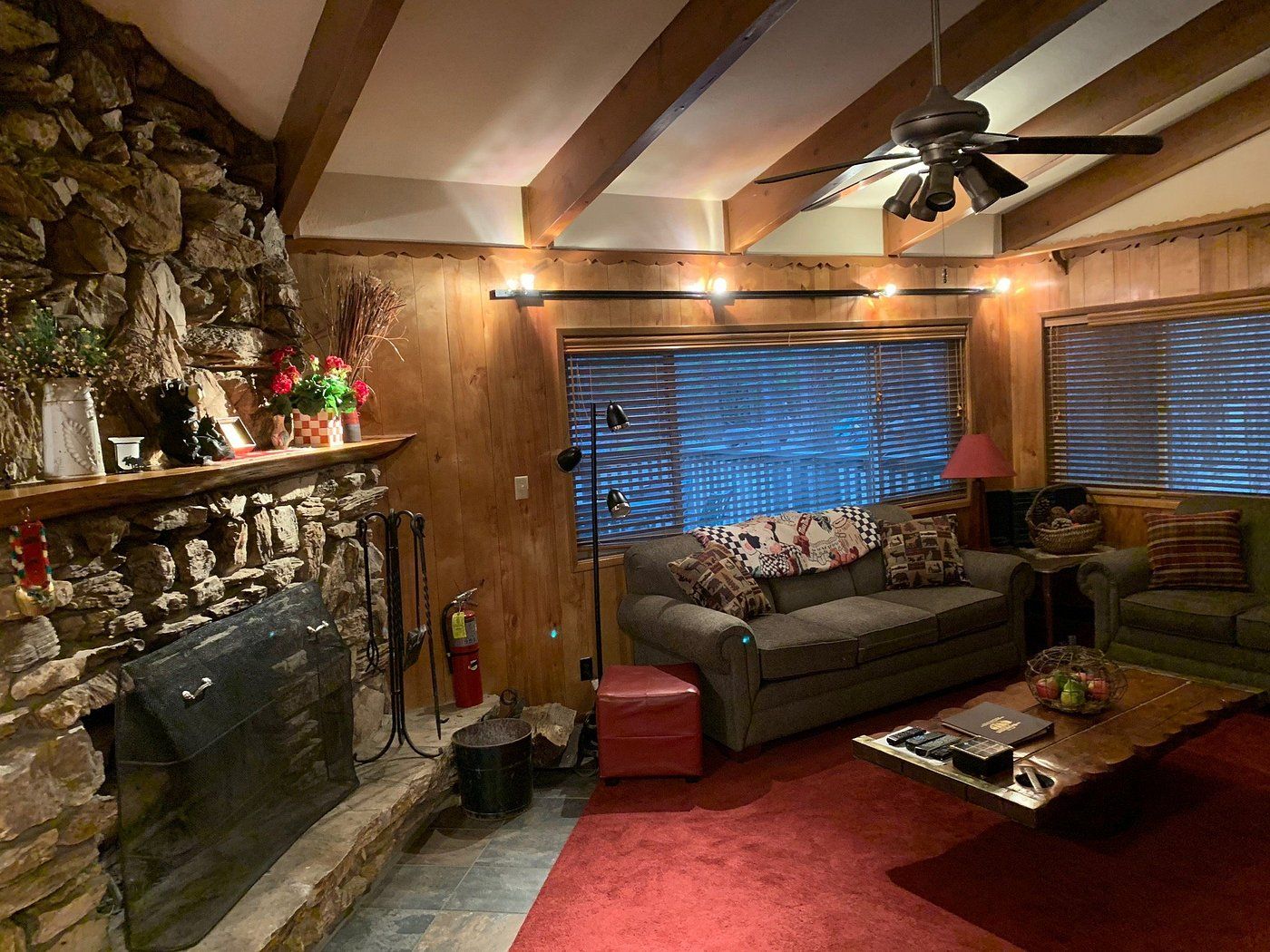 A cozy living room with a stone fireplace, wooden beams, a ceiling fan, and a window with blinds.