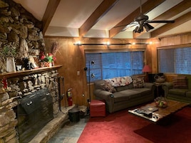 Una sala de estar acogedora con chimenea de piedra, vigas de madera, ventilador de techo y ventana con persianas.