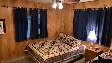 A bedroom with a wooden paneled wall, a bed with a patterned bedspread, a nightstand with a lamp, and a ceiling fan.