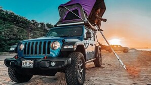 Un vehículo todoterreno azul con toldo morado sobre una playa arenosa al atardecer.
