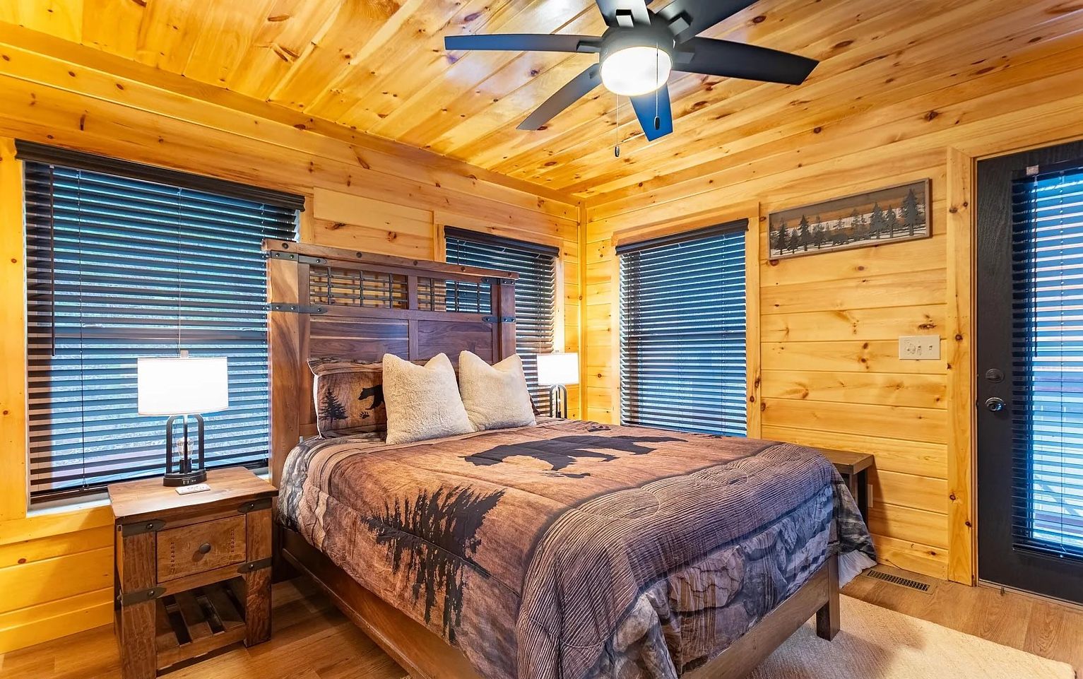 Un dormitorio de cabaña de madera con cama, mesitas de noche, ventilador de techo y ventana con persianas.
