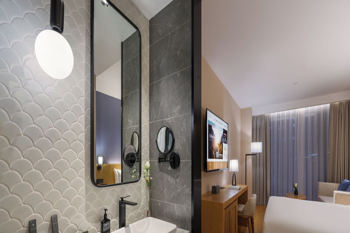 A hotel room with a large mirror, a sink, a bed, a desk, and a TV.