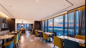 A modern restaurant with high ceilings, large windows offering a city view, and a variety of seating options.