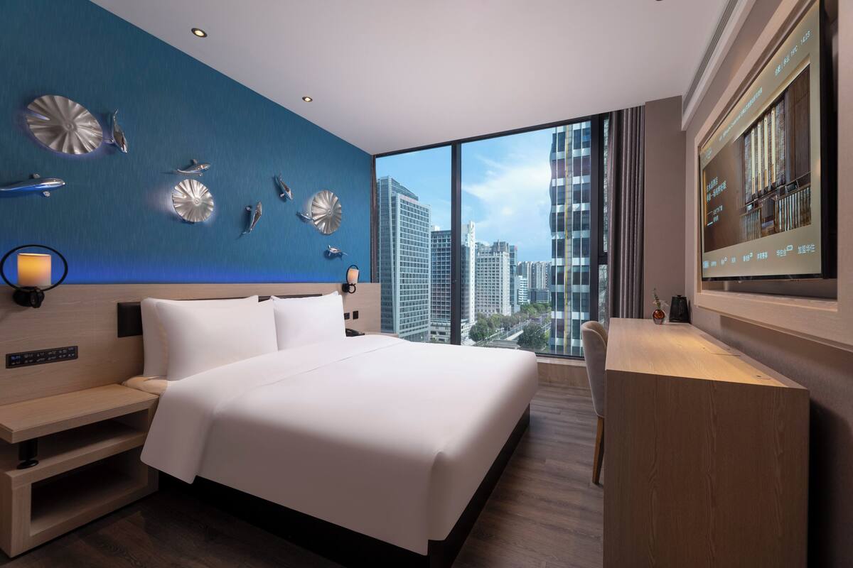A modern hotel room with a large bed, a desk, and a city view through large windows.