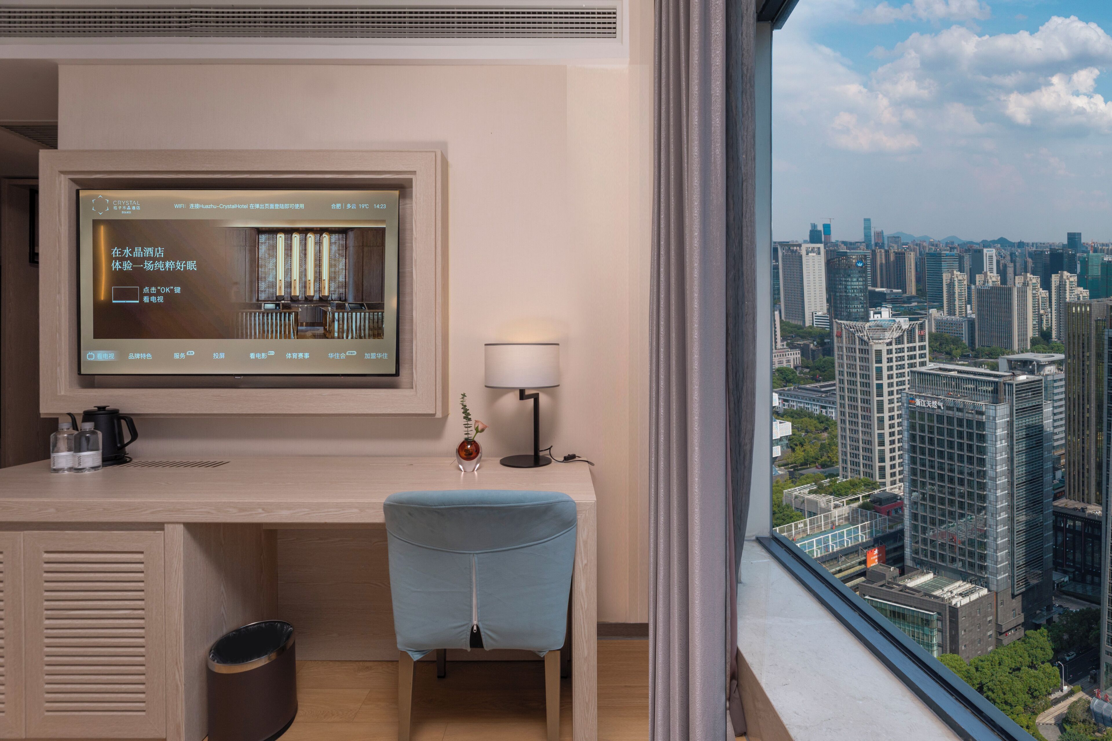 A hotel room with a large window, a desk with a computer, a chair, a trash bin, and a lamp.