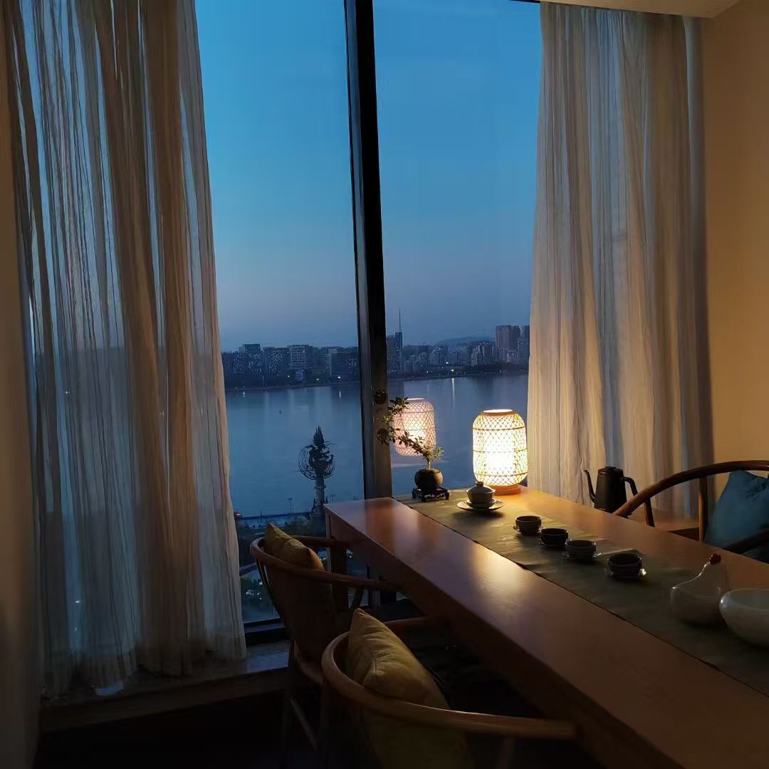 A dining area with a view of a cityscape through large windows, a table set with dishes, and a lamp providing light.
