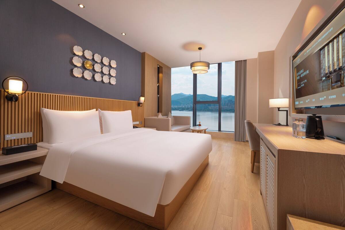 A modern hotel room with a large bed, a desk, a TV, and a view of the city.
