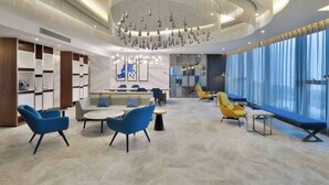 A modern hotel lobby with a central seating area, a large chandelier, and a view through large windows.