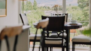 A dining area with a table and chairs, a lamp, and a view of greenery and mountains through large windows. - Beachside Luxury Suite | Pool, hottub, Sleeps 6 (Kelowna)