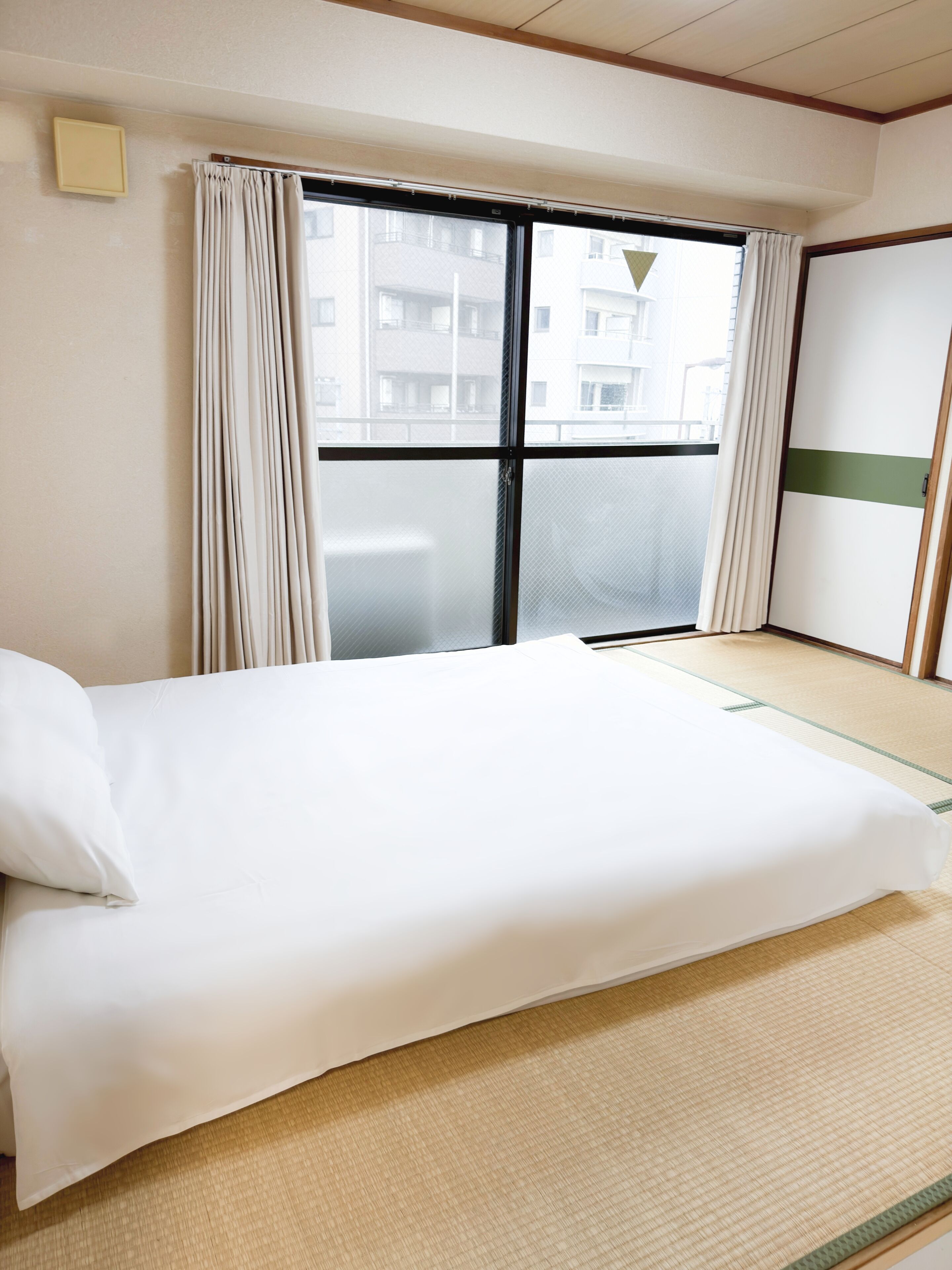 A hotel room with a bed, a window with curtains, a view of buildings outside, and a sliding door.