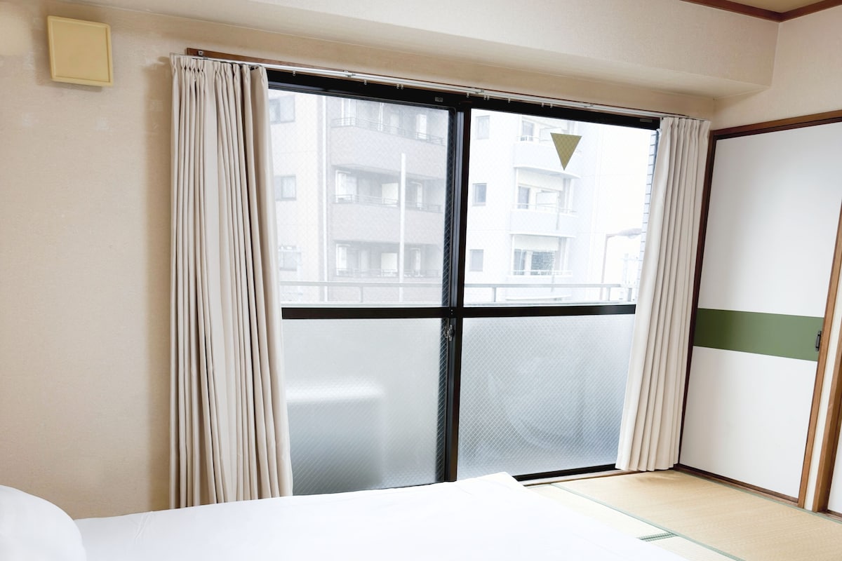 Ein Hotelzimmer mit einem Bett, einem Fenster mit Vorhängen, Blick auf Gebäude und einer Schiebetür.