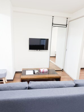 Ein modernes Wohnzimmer mit einem an der Wand befestigten Flachbildfernseher, einem Glastisch mit Gegenständen darauf und einem grauen Sofa.
