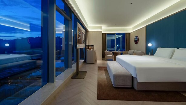 A modern hotel room with a large bed, a desk, a chair, a small table, and a large window offering a view of the cityscape. - Orange Crystal Hangzhou Tonglu Station (Hangzhou)