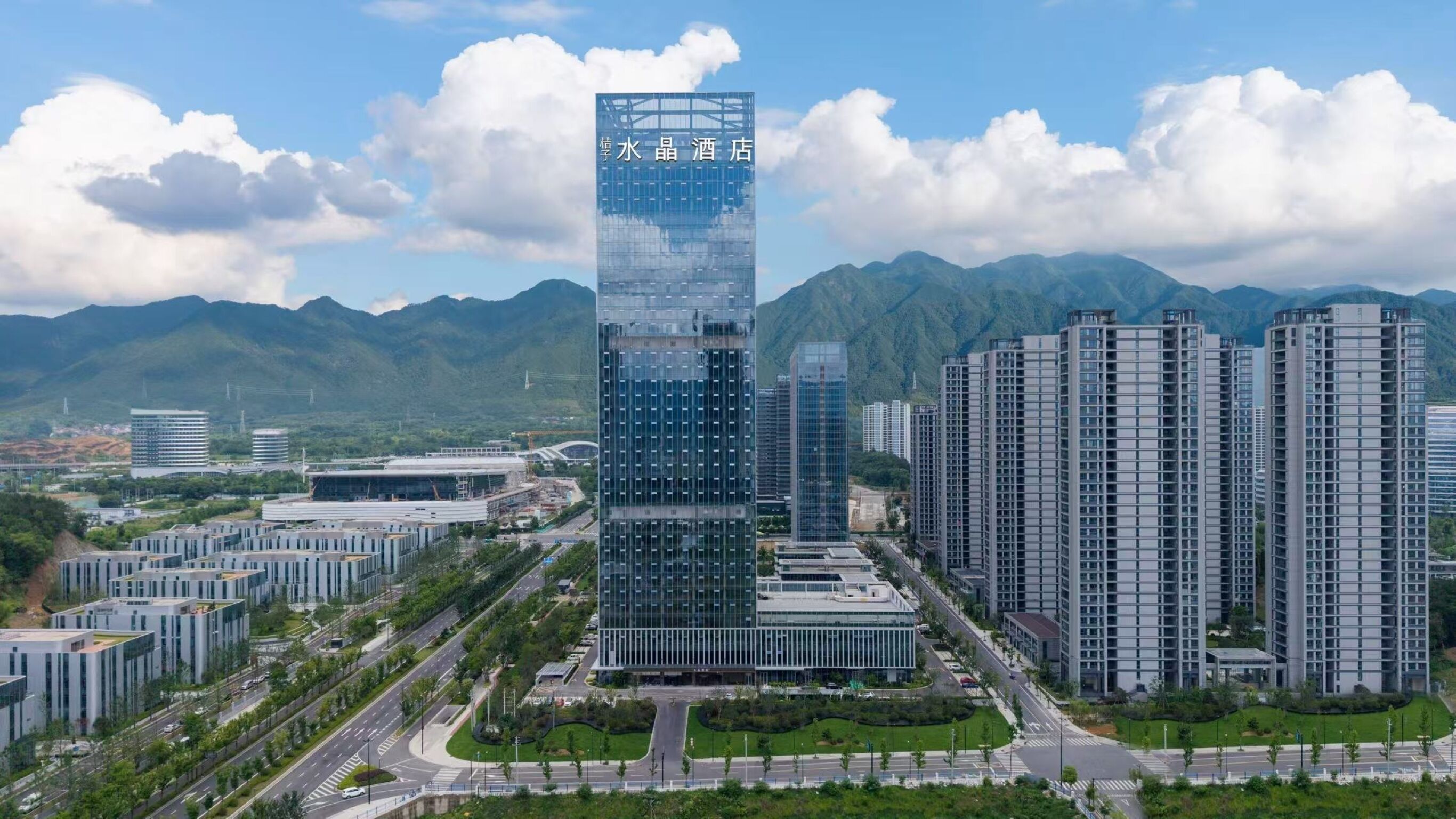 A modern hotel with a glass facade, surrounded by high-rise buildings and mountains.