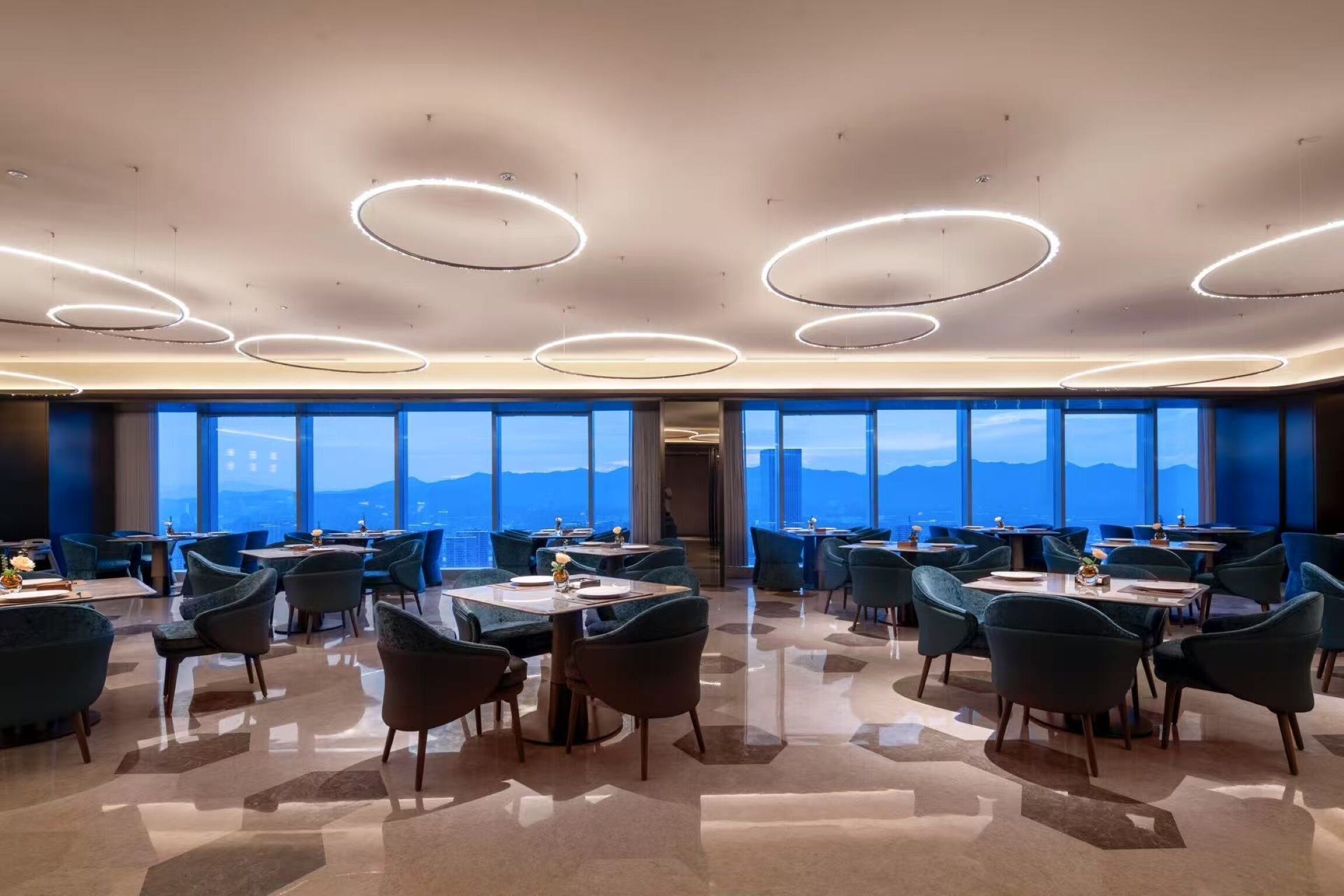 Un comedor moderno con mesas y sillas redondas, ventanas amplias con vistas y lámparas de techo circulares.