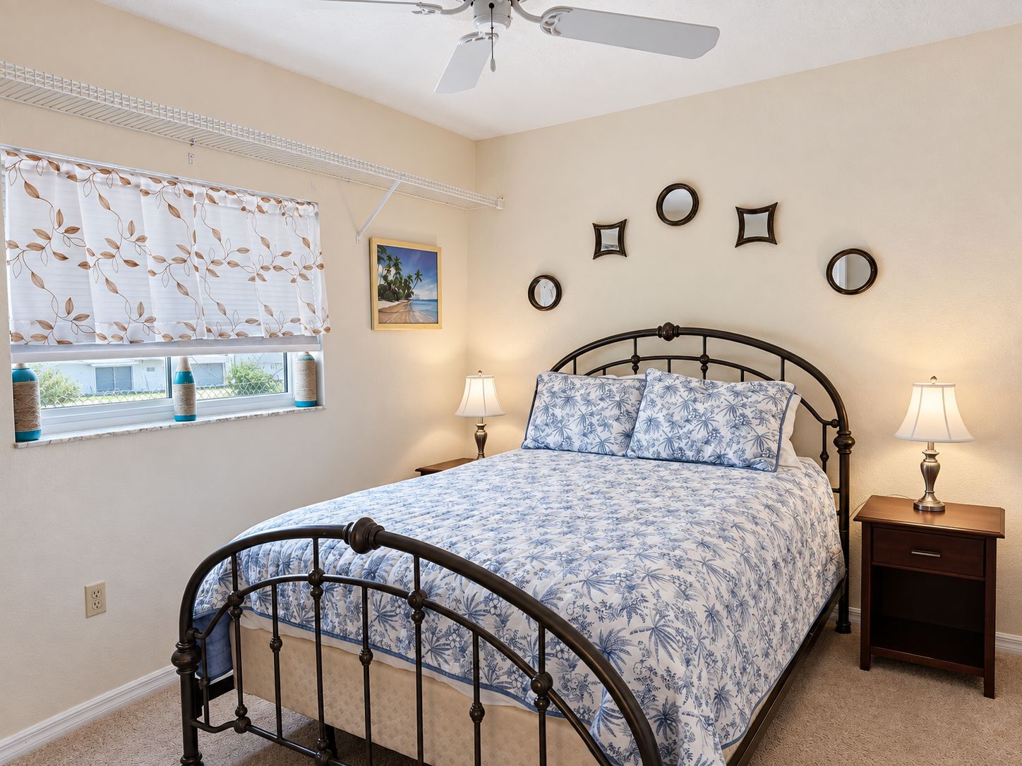 A bedroom with a bed, two bedside tables, a window with curtains, a ceiling fan, and wall-mounted mirrors.