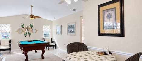 A room with a pool table, a ceiling fan, a table with a patterned tablecloth, and framed artwork on the wall.