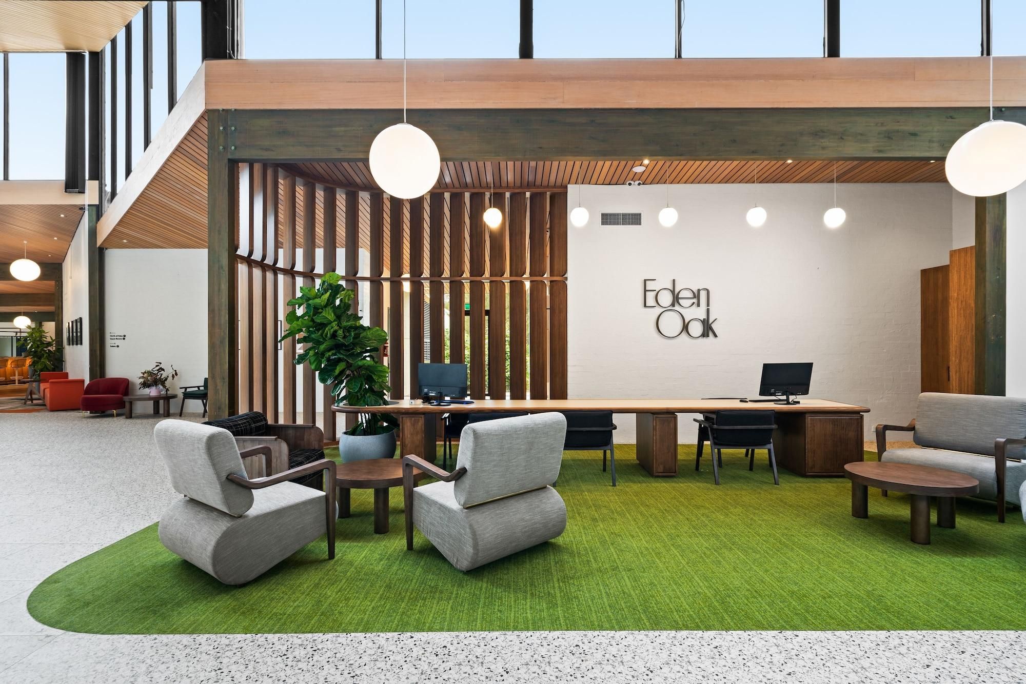 A modern office space with a wooden desk, two grey armchairs, a round table, and a potted plant.