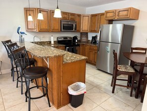 A kitchen with wooden cabinets, a granite countertop, stainless steel appliances, and bar stools. - Sunny Sands in Cocoa Beach - Partial Oceanview (Cocoa Beach)
