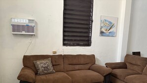 Ein Zimmer mit einer braunen Couch, einem gemusterten Kissen, einer Wand-Klimaanlage, einem Fenster mit Jalousien und einem karierten Boden.