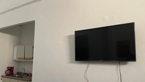 Ein Zimmer mit einem an der Wand befestigten Flachbildfernseher, einer Küchenzeile mit Schränken und einem roten Gerät, sowie einem sichtbaren Stromkabel.