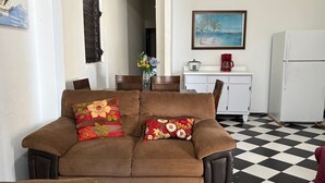 Ein Wohnzimmer mit einer Couch, einem Esstisch, Stühlen, einer Küchenzeile mit Kühlschrank und einem Bild an der Wand.