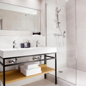 Un baño moderno con ducha envuelta en vidrio, un tocador blanco con estructura negra y un estante de madera con toallas.