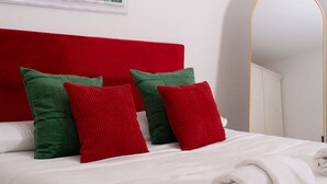 Ein Bett mit weißer Bettwäsche, roten und grünen Kissen, einem weißen Kopfteil und einer weißen Wand mit einem gerahmten Bild.