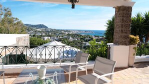 Eine Terrasse mit Meerblick, weiße Stühle, ein Tisch mit einer Vase und eine Zierensäule.