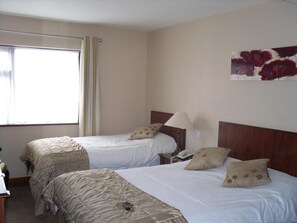 A hotel room with two beds, a window with curtains, a bedside lamp, and a floral artwork on the wall. - The Horse And Hound Inn Hotel (Ballynabola)