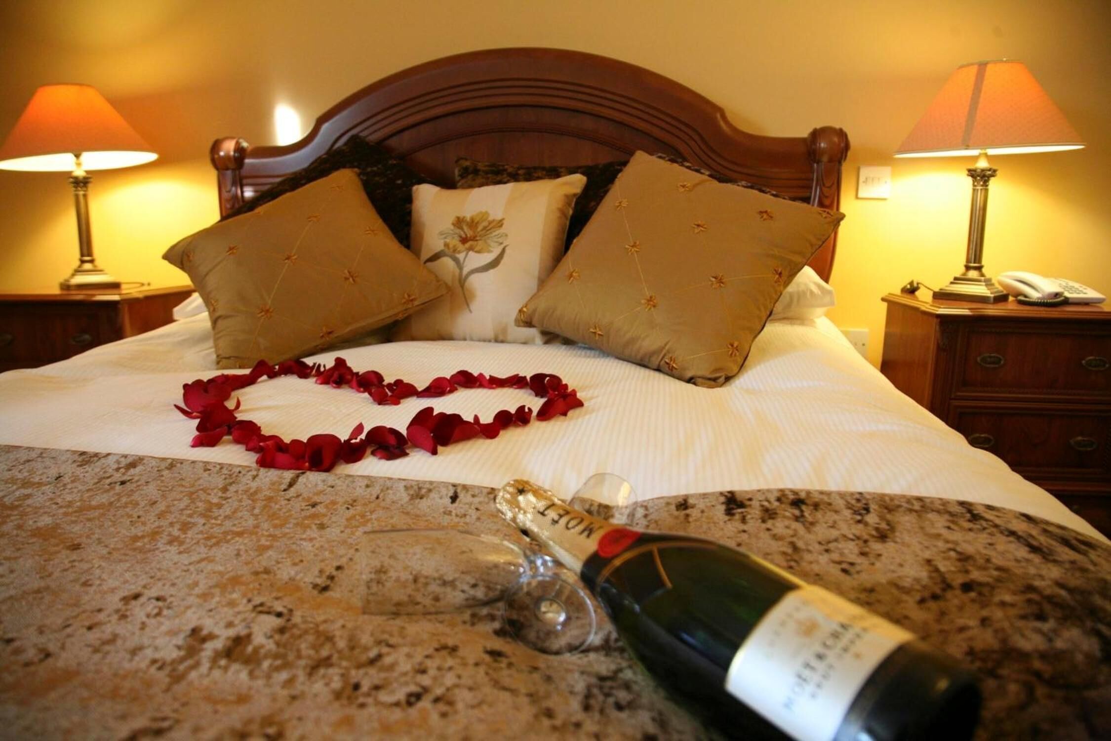 Ein Bett mit einer herzenförmigen Anordnung aus Rosenblättern, einer Champagnerflasche und zwei Lampen auf Nachttischen.