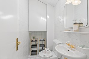 Ein Badezimmer mit weißen Fliesen, einer Toilette, einem Waschbecken mit goldfarbenen Armaturen, einem Spiegel und einem Ablagefach mit Pflegeprodukten.
