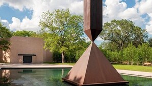 Una escultura moderna en un patio con piscina, rodeado de árboles y un edificio.