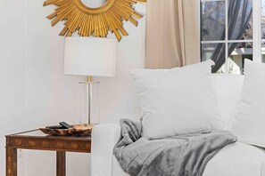 Un canapé blanc surmonté d’un plaid gris, une table d’appoint en bois avec une lampe, et un grand miroir rond doré accroché au mur.