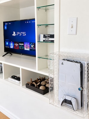 Un mueble blanco de entretenimiento con un televisor mostrando juegos de PS5, una consola PlayStation y fundas de juegos.