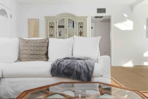 Un sofá blanco con almohadones estampados, una mesa de centro de vidrio y un aparador blanco con puertas de cristal al fondo.