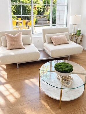 Un salon moderne avec deux canapés, une table basse en verre ornée d’une plante verte, et une vue sur un jardin grâce à de grandes fenêtres.
