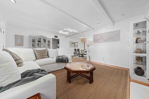 A modern living room with a white sofa, a glass coffee table, and a dining area in the background. - 4BR Cottage • Relaxed Outdoor Living Near Expo (Sacramento)